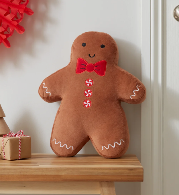 A gingerbread man plush pillow with a red bow tie and candy buttons, standing on a wooden shelf in a home setting.